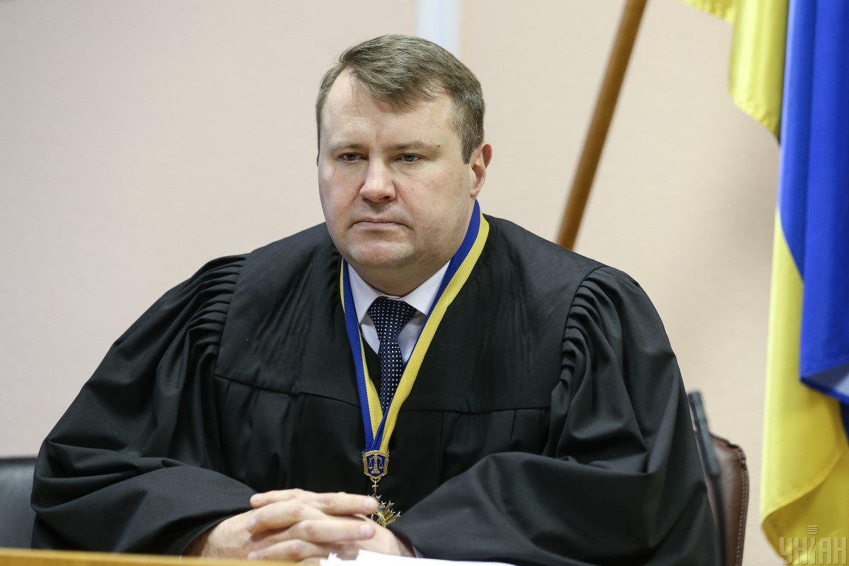 Чоловік сидить у кріслі за столом у суддівській мантії, на фоні справа - прапор України