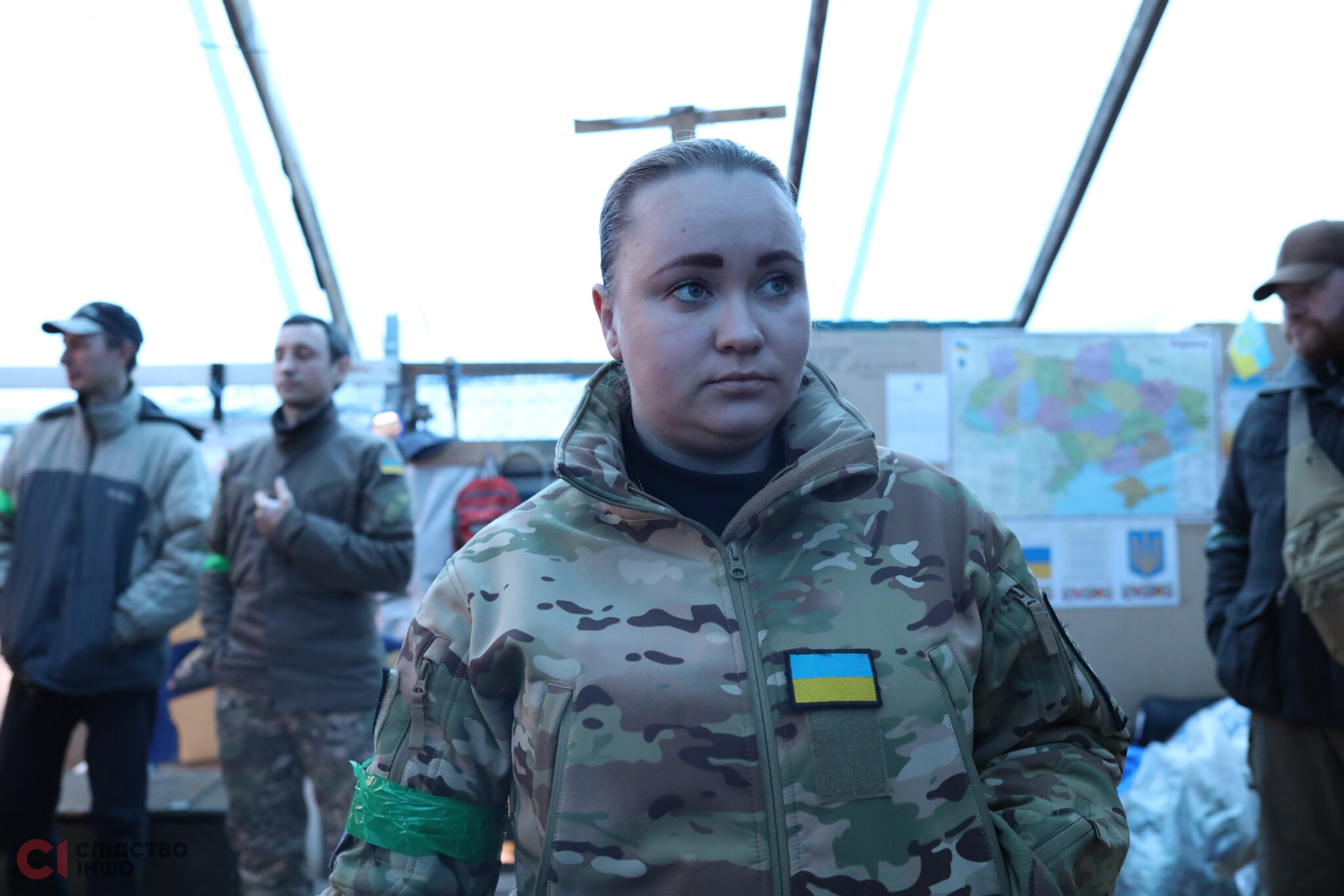 На передньому плані жінка у військовій формі з зібраним волоссям, на грудях шеврон із прапором України. Позад неї стоїть троє чоловіків: двоє - ліворуч і один - праворуч