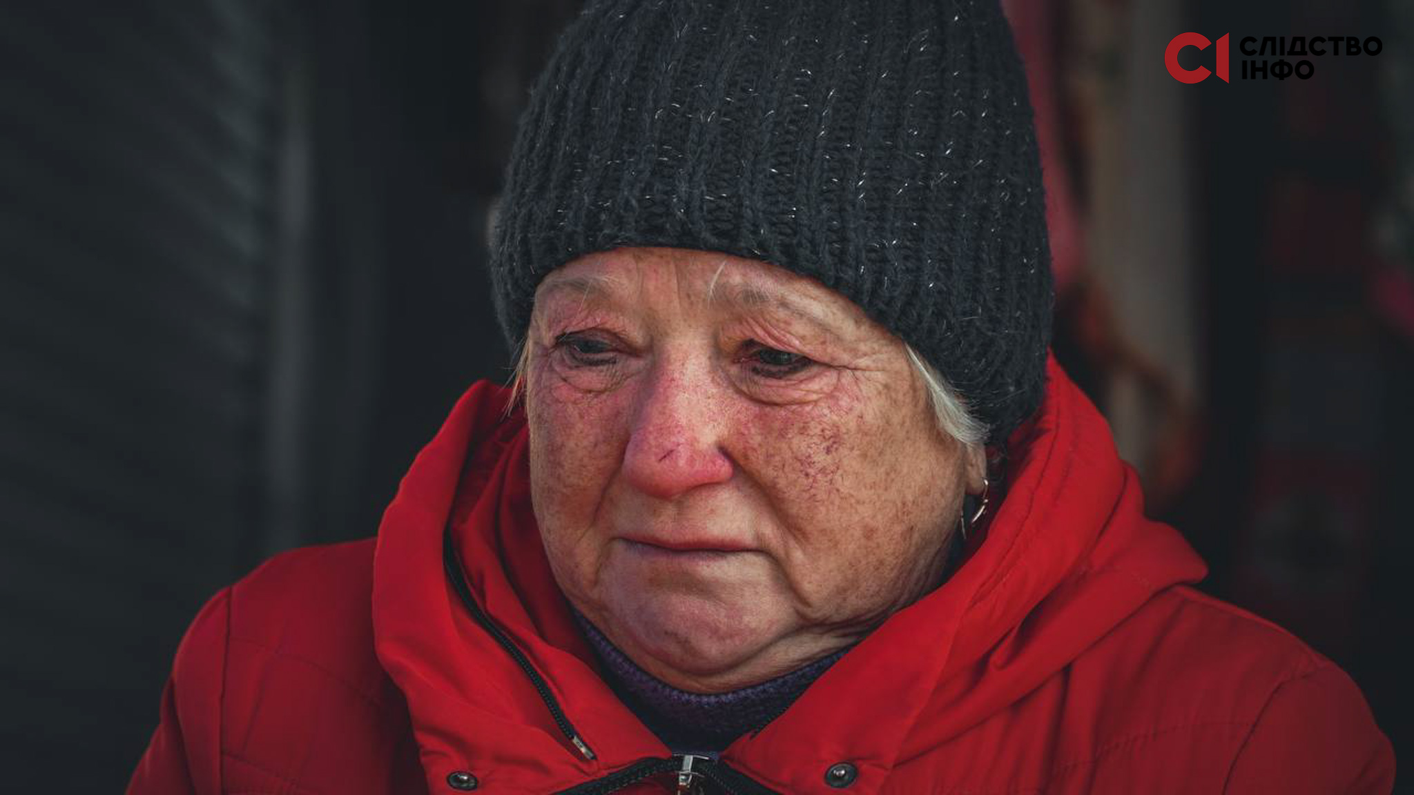 Заплакана старша жінка у чорній шапці й червоній куртці