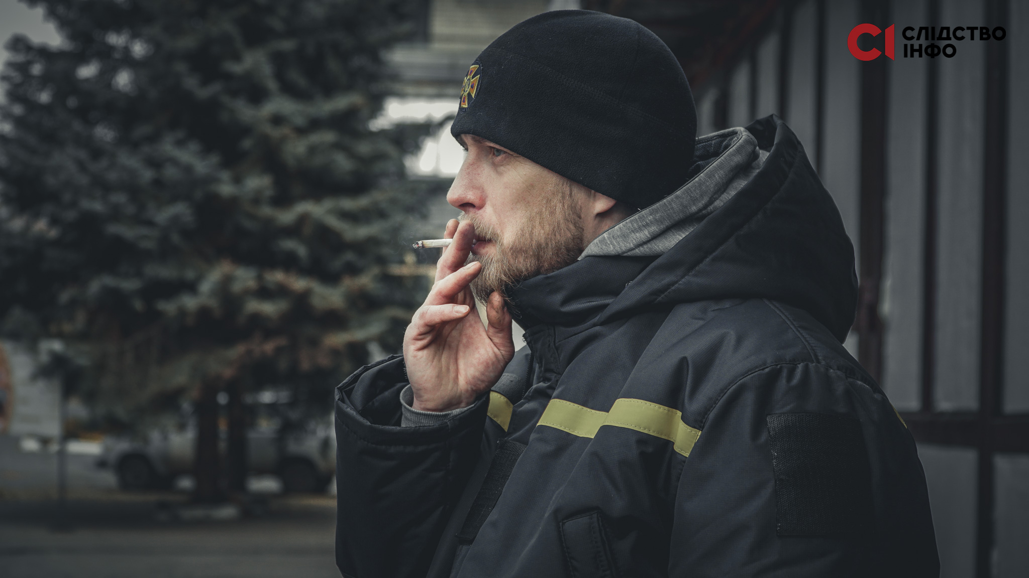 Чоловік стоїть в профіль на вулиці й курить сигарету. На ньому чорна шапка. На фоні - ялинки