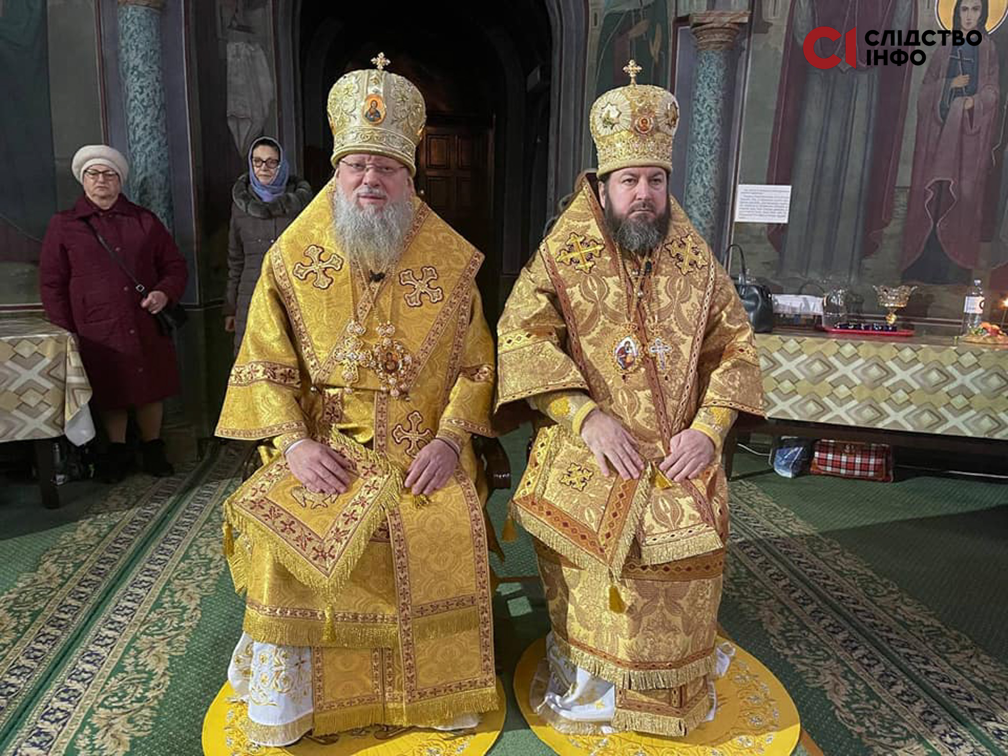 На фото сидять два священника РПЦ в Україні у золотих святкових рясах, позад них зліва стоять дві жінки в окулярах 