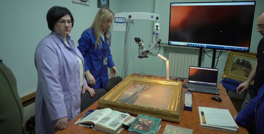 На фото зліва біля столу стоять дві жінки, на столі лежить картина Айвазовського в позолоченому обрамленні. Справа на стіні висить велика плазма, біля якої стоїть чоловік в окулярах