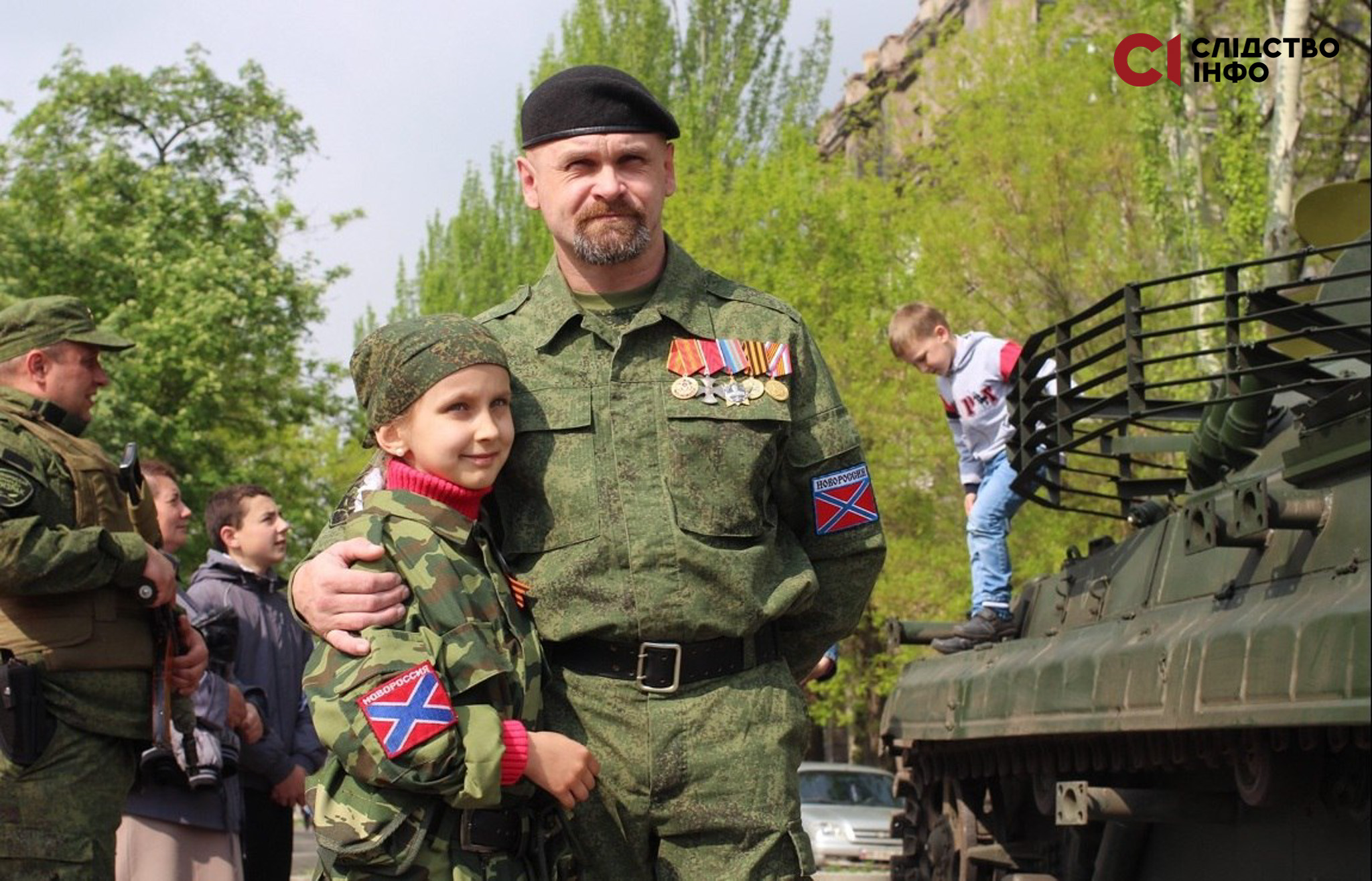 На фото чоловік у російській військовій формі обіймає маленьку дівчинку у військовій формі. Праворуч стоїть танк, на якому знаходиться маленький хлопчик. На фото є інший чоловік у формі з автоматом, який дивиться у бік хлопчика. 