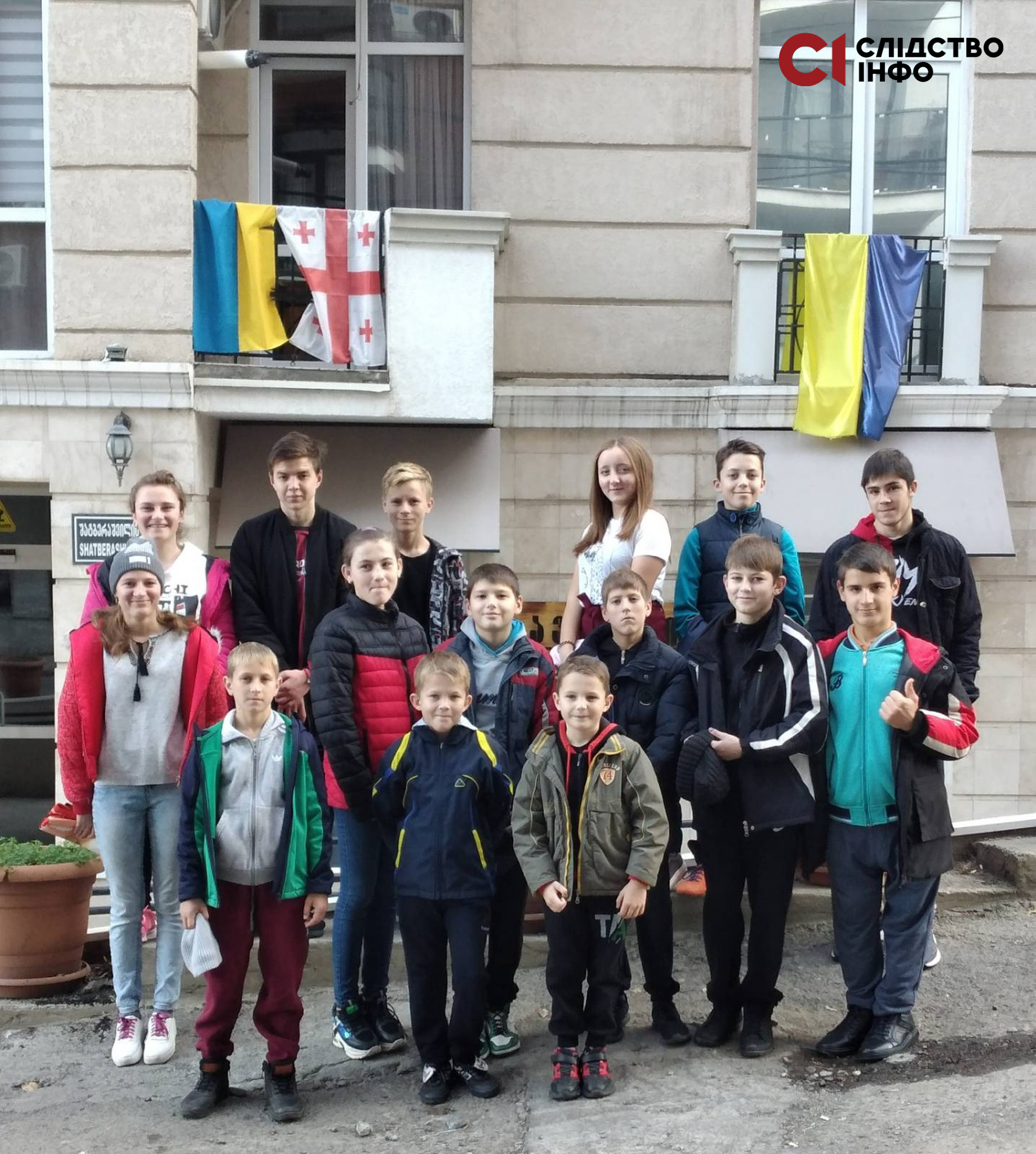 На фото 15 українських дітей, яких вдалося вивезти з Росії в Грузію. Діти стоять на вулиці, позад них будинок з двома великими вікнами, на яких висять прапори України та Грузії 